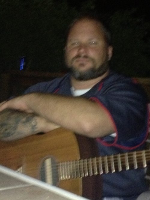 Ran Kime sitting with an acoustic guitar, reflecting the quiet moments of writing and rehearsal behind brothers KIME recordings.