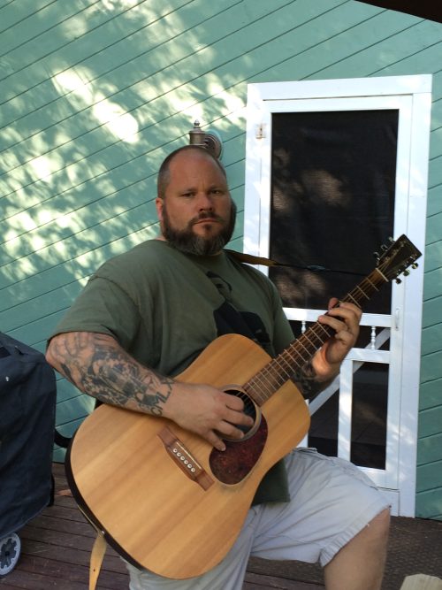 Ran Kime of brothers KIME playing acoustic guitar on a sunlit deck, capturing the raw, stripped-down songwriting moments behind the band’s sound.